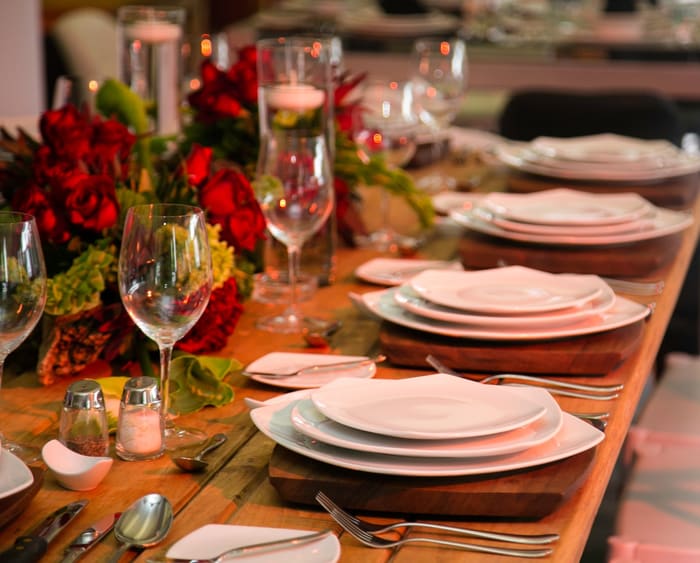 Mesa larga de madera con vajilla blanca y centro de flores rojas